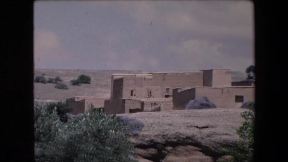1971 Algeria 8mm frame showing a distant village edge with earthen flat-roofed buildings, scrub vegetation, dry open ground, and tiny human figures near the walls.