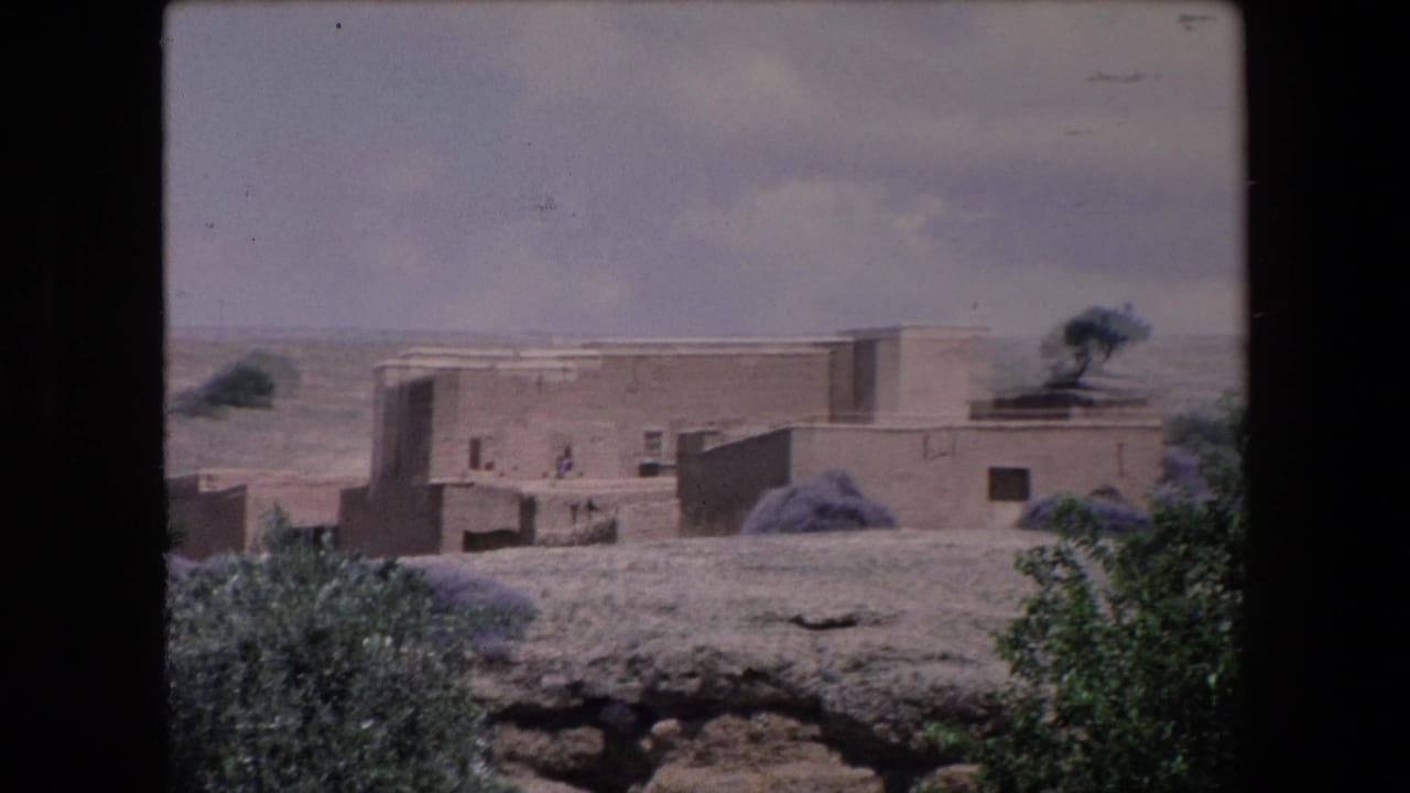 1971 Algeria 8mm frame showing low adobe buildings with flat roofs, sparse shrubs, dry terrain, and small distant figures under a cloudy sky.