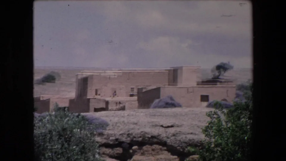 1971 Algeria 8mm frame showing low adobe buildings with flat roofs, sparse shrubs, dry terrain, and small distant figures under a cloudy sky.