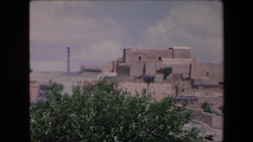 1971 Algeria 8mm frame showing a larger adobe building cluster, foreground greenery, dry village terrain, cloudy sky, and a distant utility pole.