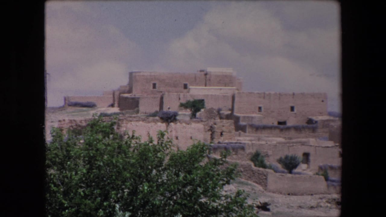 1971 Algeria 8mm frame showing earthen flat-roofed buildings on a dry hillside with shrubs in the foreground and cloud cover above.