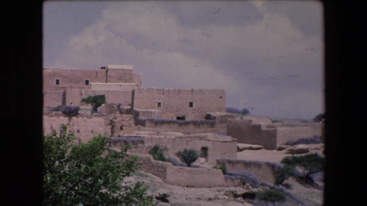 Earthen village structures and sparse shrubs across a dry slope.
