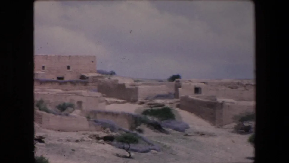 1971 Algeria 8mm frame looking across low mud-brick walls, sandy terrain, scattered shrubs, and distant earthen buildings.