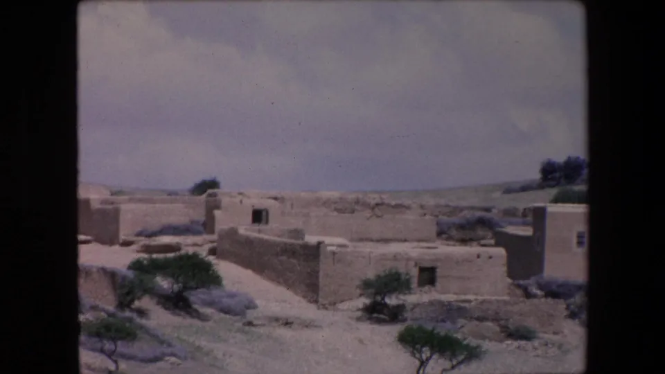 1971 Algeria 8mm frame showing mud-brick village walls, small desert trees, dry open ground, and flat-roofed structures in the background.