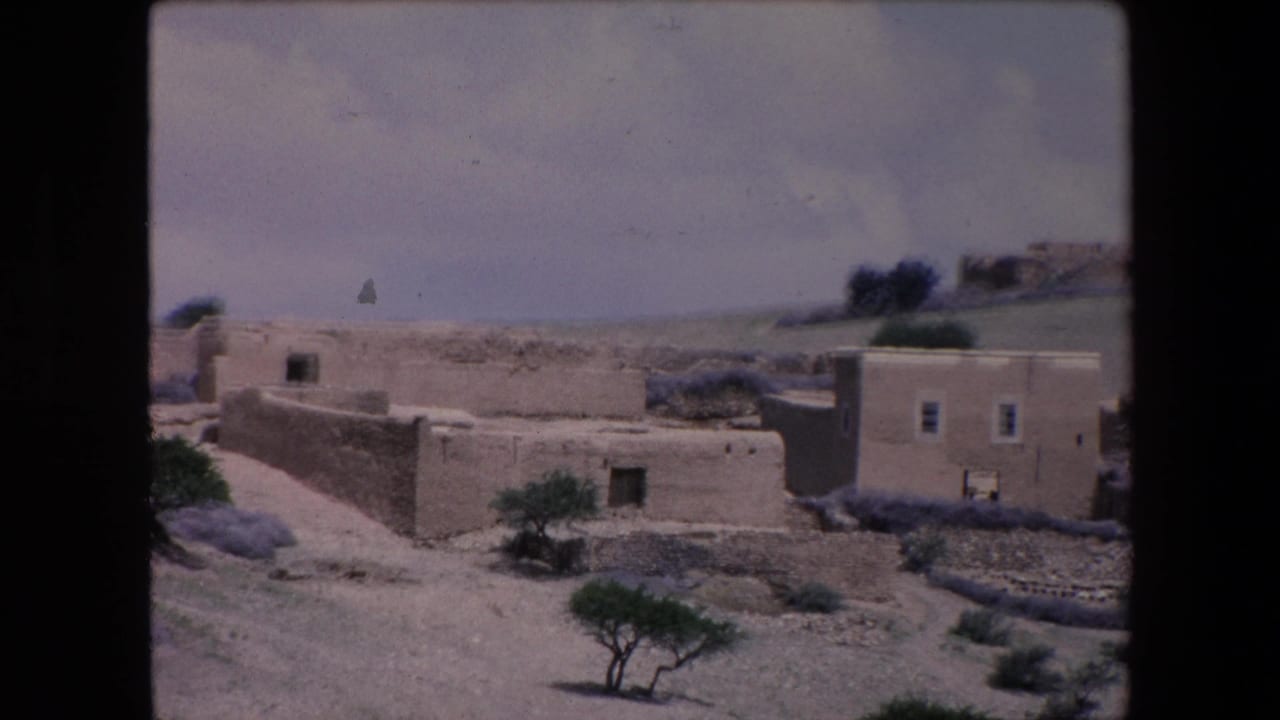1971 Algeria home-movie frame showing low stone and adobe houses on dry open ground with sparse shrubs, small trees, and distant hills.