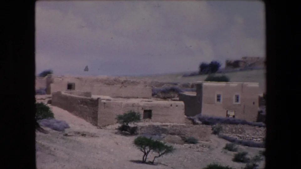 1971 Algeria home-movie frame showing low stone and adobe houses on dry open ground with sparse shrubs, small trees, and distant hills.