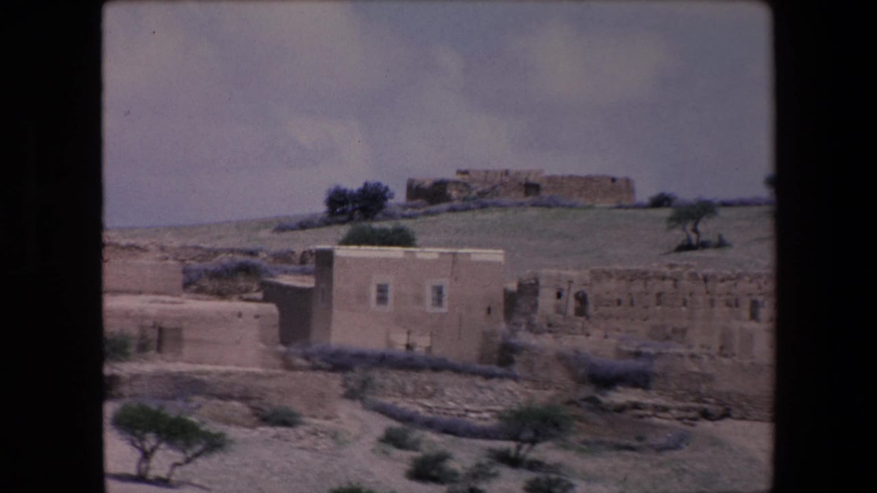 1971 Algeria frame with a small cluster of low rural houses, rough walls, sparse vegetation, and a pale sky in an arid setting.
