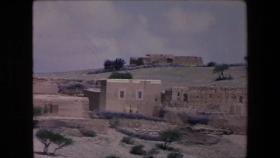 1971 Algeria frame with a small cluster of low rural houses, rough walls, sparse vegetation, and a pale sky in an arid setting.