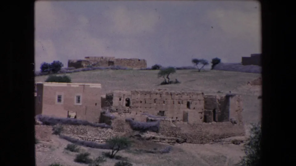 1971 Algeria home-movie frame showing a light adobe building beside a weathered stone structure, with shrubs and small trees in the foreground.