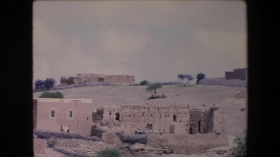 1971 Algeria frame showing weathered rural buildings on dry terrain, with a light-colored house, rough stone walls, sparse trees, and a pale sky.