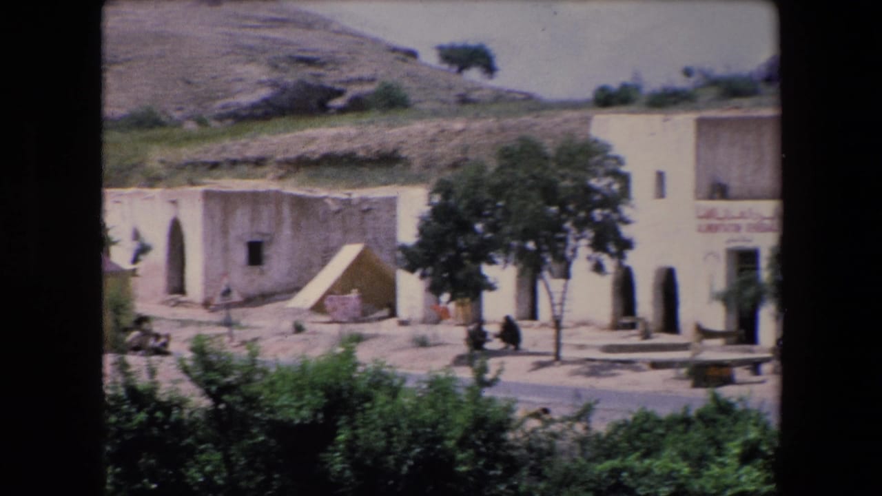 Distant village storefront with white buildings, trees, hillside, and seated people.