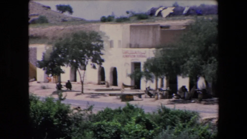 1971 Algeria village view with a bilingual general store sign on a pale building, arched openings, shade trees, benches or low tables, and people gathered near the facade.