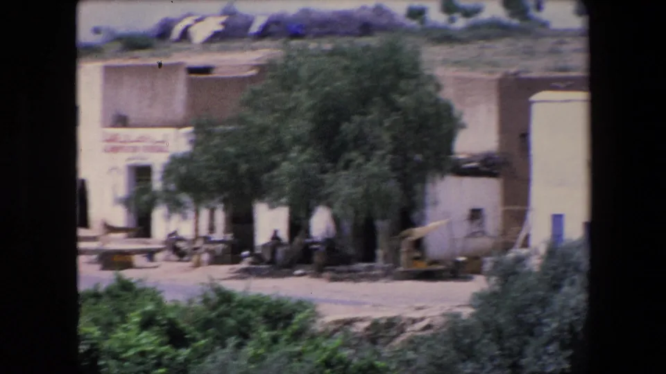 1971 Algeria frame from a moving 8mm shot, with the general store building softened by motion, a large tree in front, hillside behind, and figures seated near the shaded exterior.