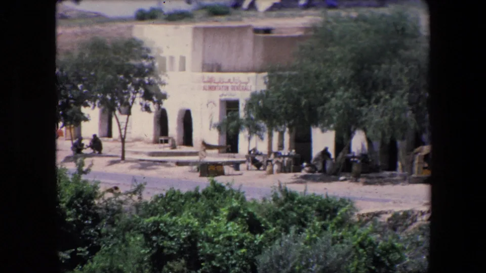 1971 Algeria home-movie still showing a quiet storefront with Arabic and French signage, arched entries, trees casting shade, low outdoor seating, foreground greenery, and distant seated people.