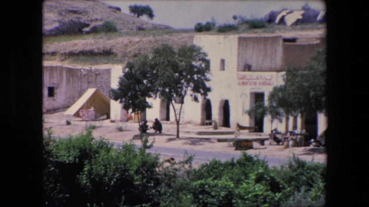 1971 Algeria 8mm frame showing a pale building with Arabic lettering beside trees, shrubs, and a dry hillside near the river area.