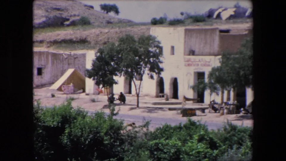 1971 Algeria 8mm frame showing a pale building with Arabic lettering beside trees, shrubs, and a dry hillside near the river area.