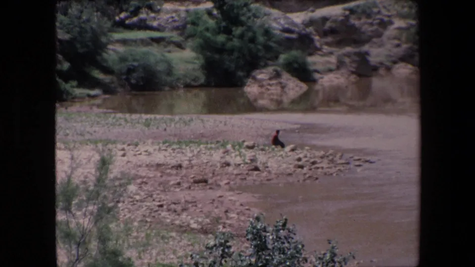 1971 Algeria 8mm frame with a lone dark-clothed person seated on a rocky riverbank across from shrubs, boulders, and arid terrain.