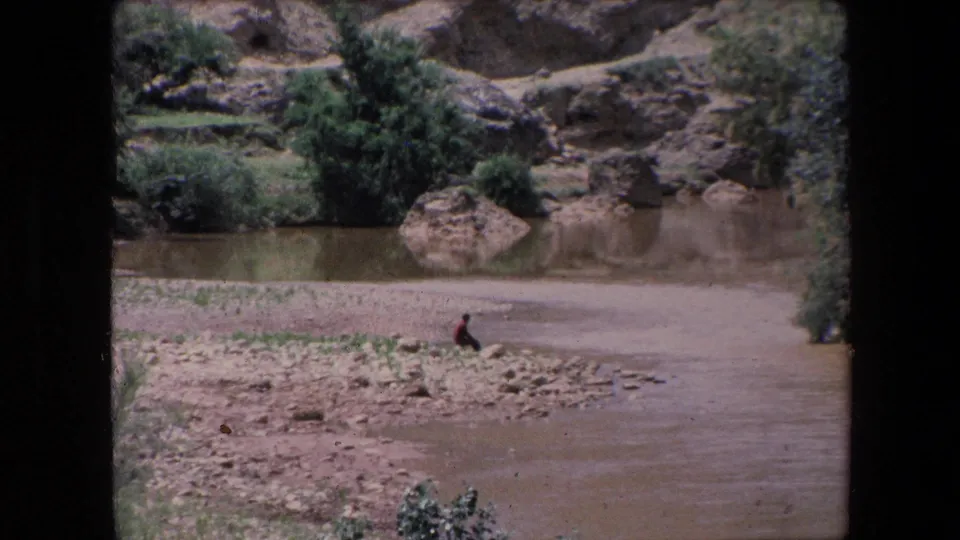 1971 Algeria 8mm frame of a lone person sitting close to the muddy river, with rocks, shrubs, a large boulder, and arid slopes in the background.