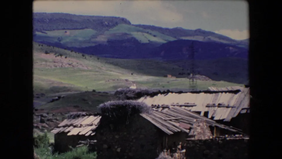 A rural Algerian hillside landscape shows stone buildings with weathered roofs in the foreground and green rolling hills beyond.