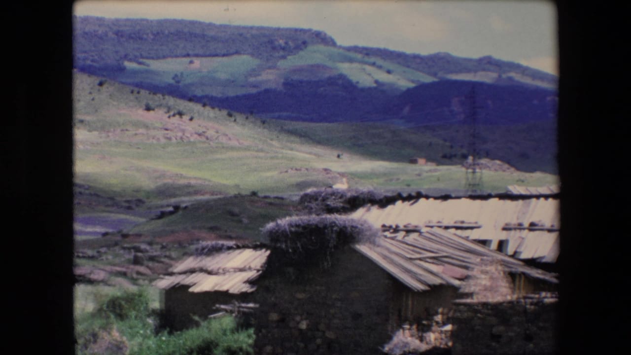 1971 Algeria 8mm frame showing weathered stone and wooden buildings with patched corrugated roofs below green hills and distant mountains.
