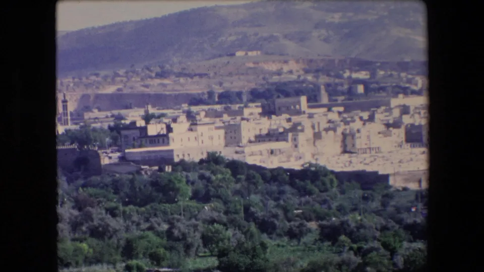 1971 Algeria 8mm frame showing a sunlit townscape, low-rise buildings, foreground greenery, and broad hills behind the settlement.