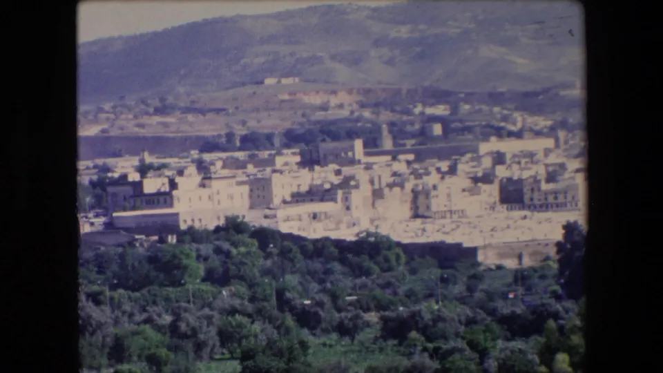 1971 Algeria 8mm frame presenting a distant town, dense green foreground vegetation, pale buildings, and rolling hills in the background.