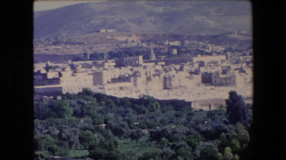 1971 Algeria 8mm frame showing an elevated view across green foreground trees toward a pale historic town and dry hills in the distance.