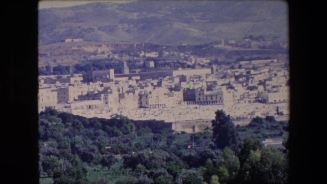 Panoramic town view with a tower rising among pale buildings and hills behind.