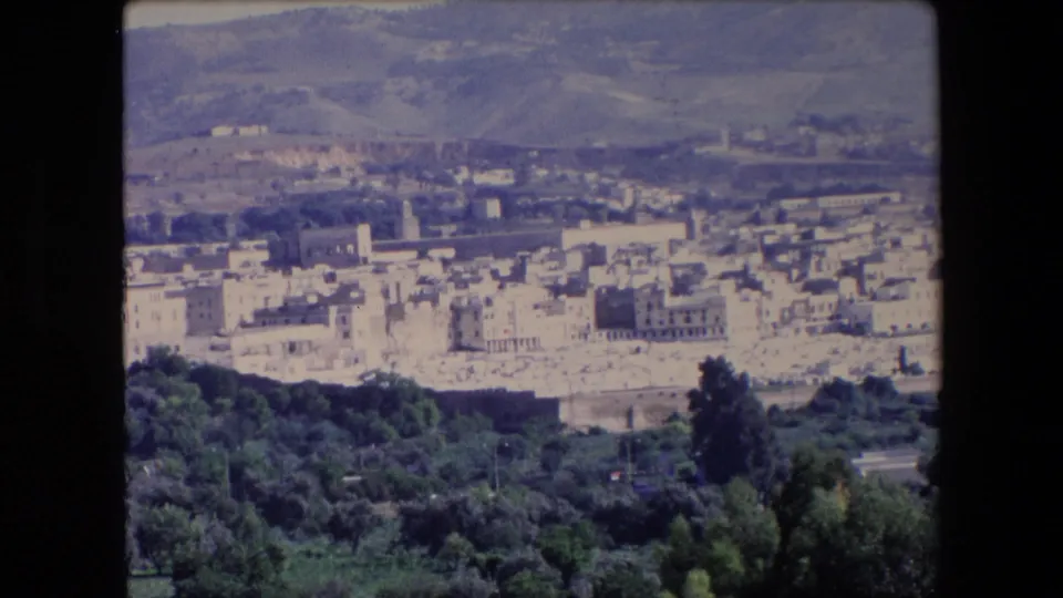 1971 Algeria 8mm frame of a dense pale townscape below hills, with a tower or minaret visible among the buildings and trees below the overlook.