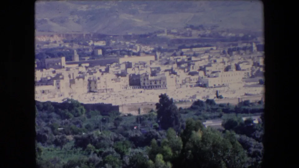 1971 Algeria 8mm frame showing the camera panning across white and beige town buildings, foreground vegetation, and hazy hills beyond.