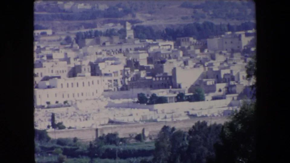 1971 Algeria 8mm frame showing a closer elevated view of dense pale buildings, a tower or minaret-like vertical structure, foreground trees, and a road at the lower edge.