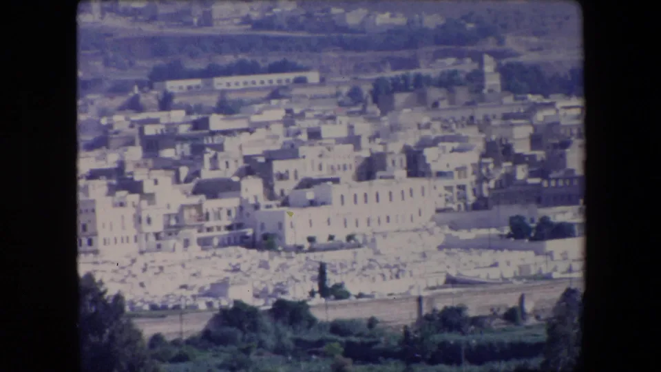 1971 Algeria 8mm frame of a hillside townscape with light buildings, a possible tower form, foreground trees, a lower road, and hazy hills beyond.