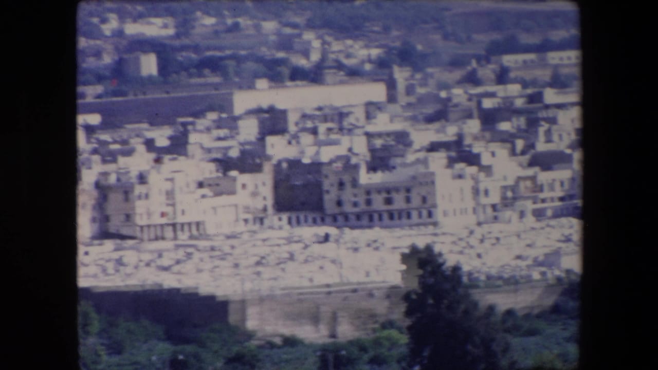 1971 Algeria frame with tightly packed white buildings, a dark stone wall, pale cemetery markers, and hillside development in the distance.