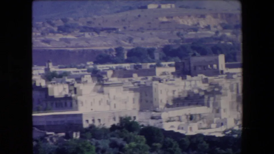 1971 Algeria frame from a panning view across white town buildings, dark trees, hillside terrain, and a cemetery-like area near the lower edge.