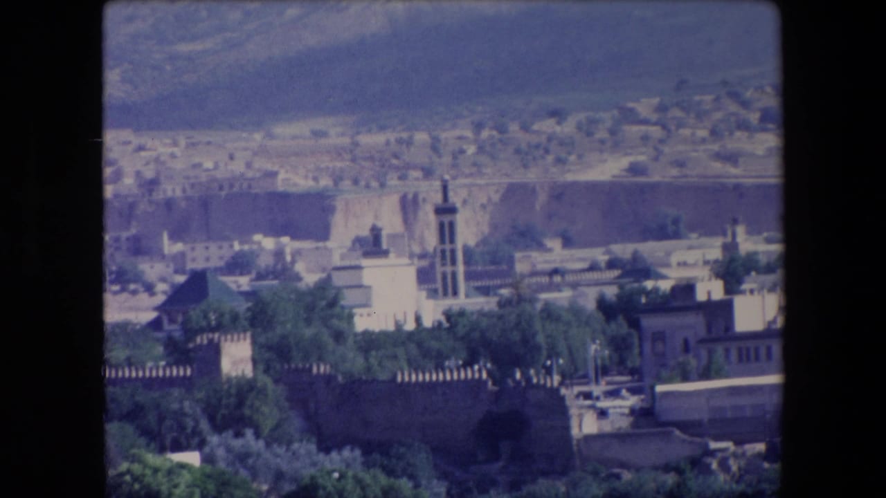 1971 Algeria 8mm frame with a tower rising above white buildings, crenellated walls, dense greenery, and a dry hillside beyond the city.