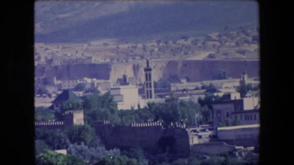 1971 Algeria 8mm frame with a tower rising above white buildings, crenellated walls, dense greenery, and a dry hillside beyond the city.