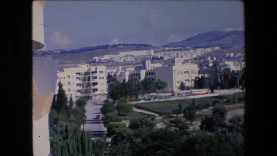 1971 Algeria 8mm frame looking across apartment blocks, a road, gardens, tall trees, and hills from an elevated visitor viewpoint.