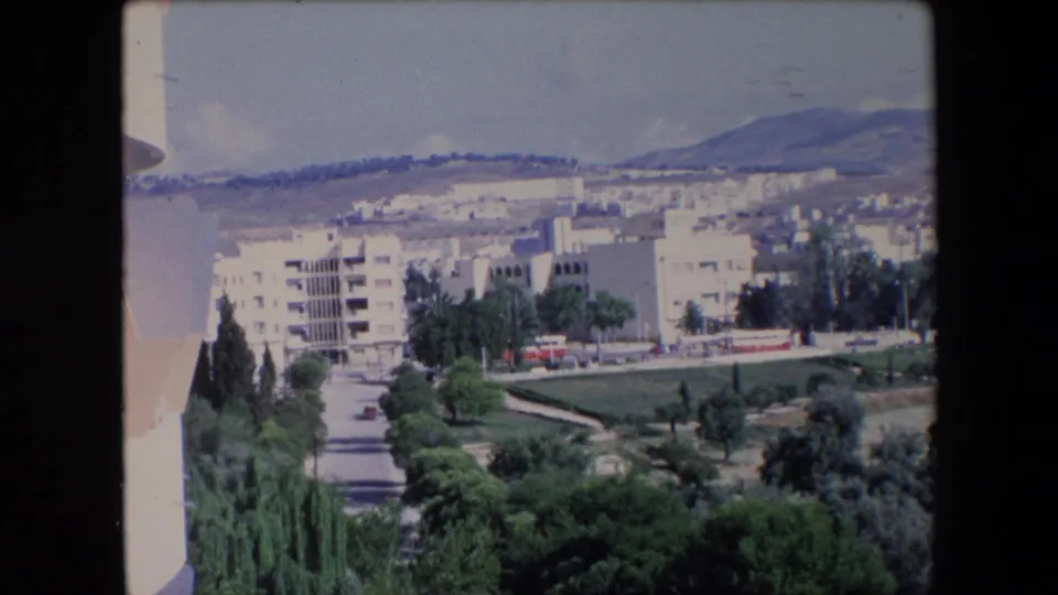 1971 Algeria 8mm frame showing pale apartment buildings, green landscaped areas, roadside trees, and distant hills under a bright sky.