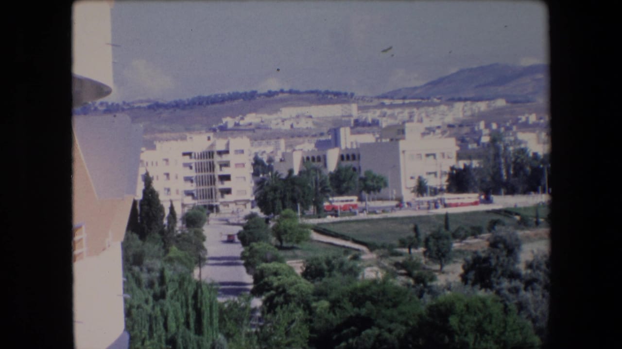 1971 Algerian 8mm frame showing a sunlit hillside town with pale apartment blocks, green trees, a road, and distant mountains under a blue sky.