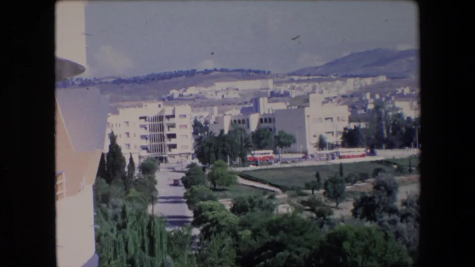 1971 Algerian 8mm frame showing a sunlit hillside town with pale apartment blocks, green trees, a road, and distant mountains under a blue sky.