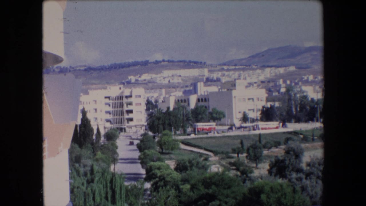 1971 Algerian home-movie frame from an elevated viewpoint, with white apartment buildings, a red bus on the road, trees, and mountains in the background.