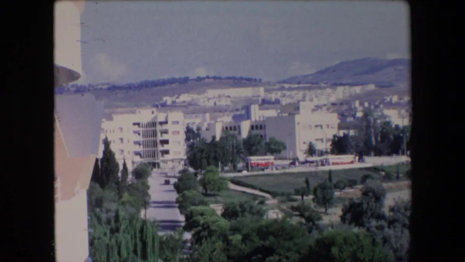 1971 Algerian home-movie frame from an elevated viewpoint, with white apartment buildings, a red bus on the road, trees, and mountains in the background.