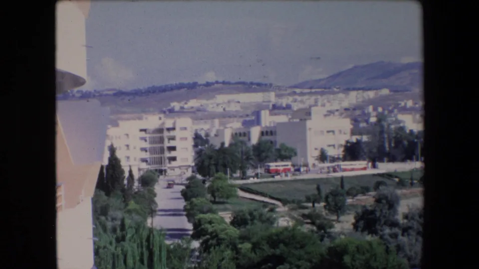1971 Algeria 8mm frame showing a repeated elevated view of pale residential blocks, a tree-lined road, foreground greenery, and hills beyond the town.