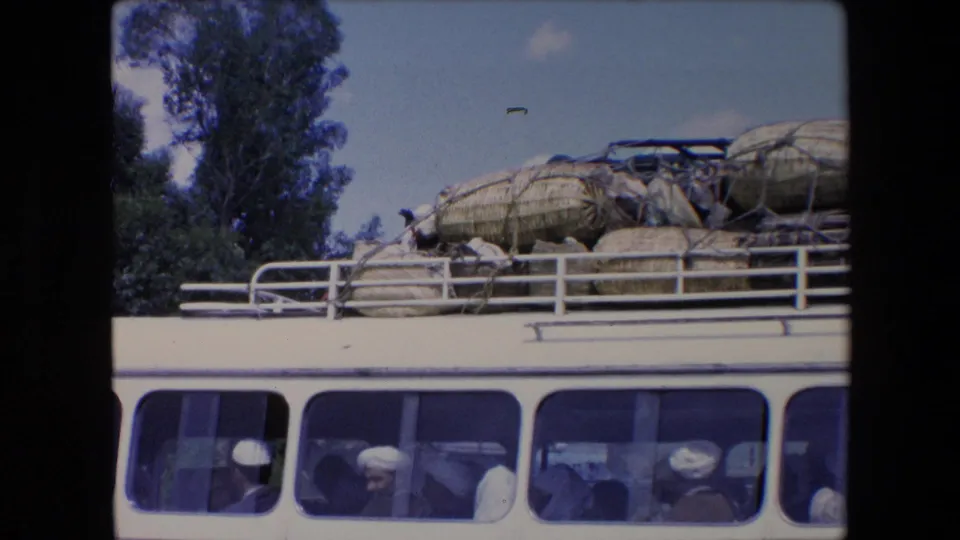 The white van passes with passengers visible through the windows while woven baskets and animals remain tied on the roof in 1971 Algeria footage.