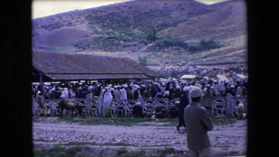 1971 Algeria home-movie frame showing a rural market or gathering beside a low tiled-roof building, with a hillside behind and a man standing in the foreground.