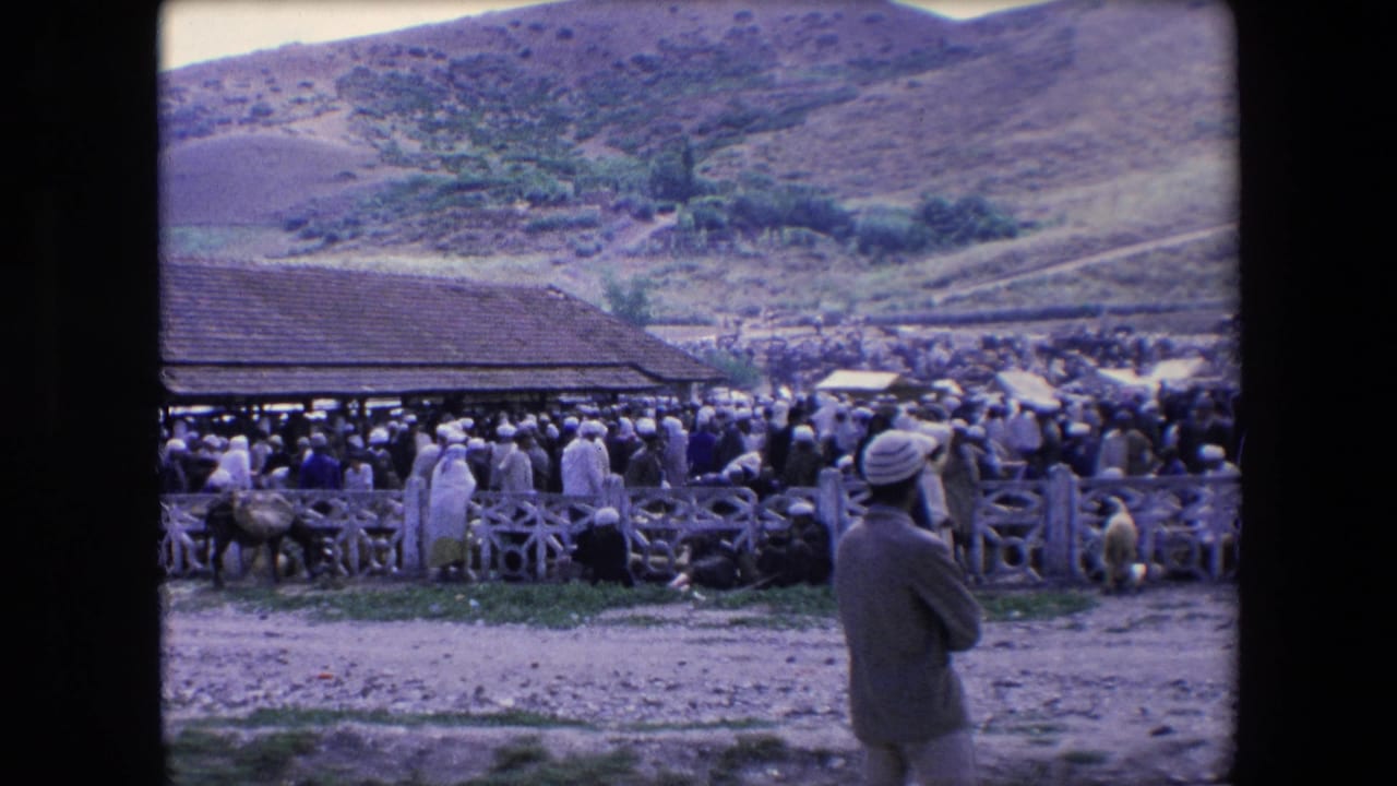 1971 Algeria 8mm frame showing a large outdoor crowd near wooden fencing and a tiled-roof structure, with a turbaned figure in the foreground and hills beyond.