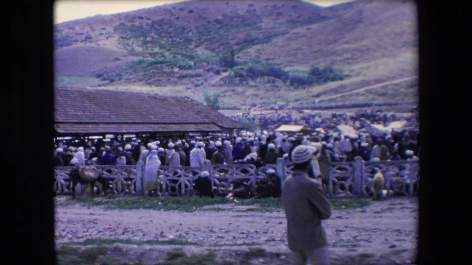 1971 Algeria 8mm frame showing a large outdoor crowd near wooden fencing and a tiled-roof structure, with a turbaned figure in the foreground and hills beyond.