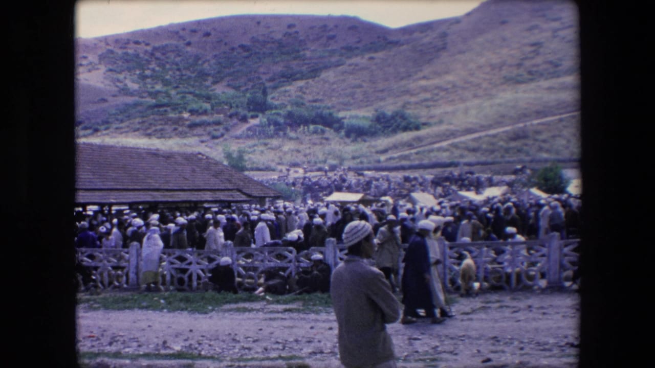Large crowd stands near a fenced market area with hills in the background.