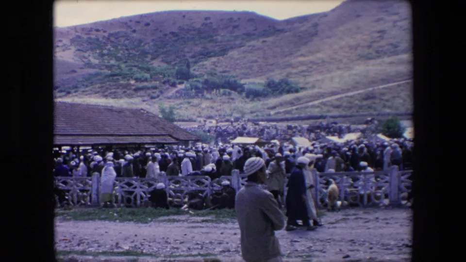1971 Algeria 8mm frame of a crowded outdoor gathering beside a rustic fence and roofed structure, with robed figures and animals visible against a mountainous backdrop.