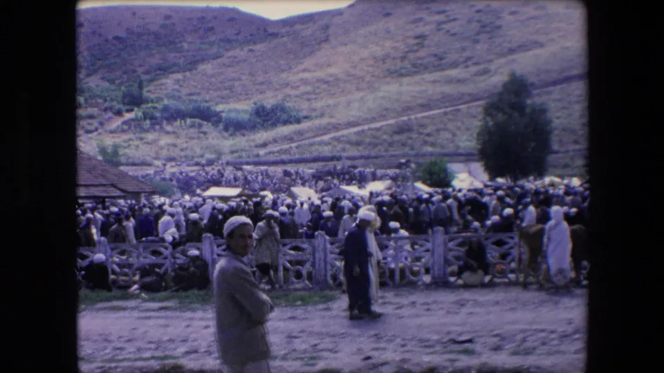 1971 Algeria 8mm frame showing a dense outdoor gathering with people in robes and turbans, wooden fencing, dusty ground, a roof edge, and rolling hills behind.